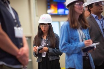 An Asian woman with long dark hair writes in a reporter's notebook while wearing a hard hat and safety glasses.