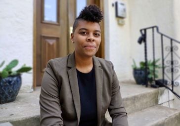 Photo of young black woman wearing black shirt and brown suit jacket sitting on steps.