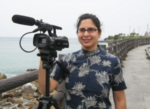 Photo of woman with black hear and glasses standing behind a video camera. There is a fence and coastal water in the background.